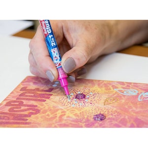 Op de afbeelding: Een hand die een blauw-witte Rolling 2-in-1 lijm pen met een roze punt vasthoudt, lijm aanbrengend op een roze en oranje patroon oppervlak. Op het oppervlak staat het woord "INSPIRE".