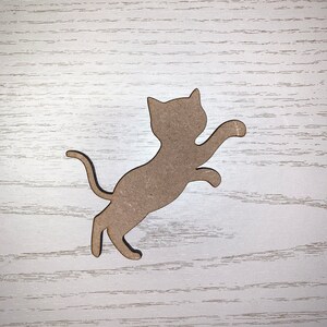 May include: A light brown cat-shaped wooden cutout, mid-jump with one paw extended. The cat has a curved tail and pointed ears. The cutout is set against a light wood-grain background.