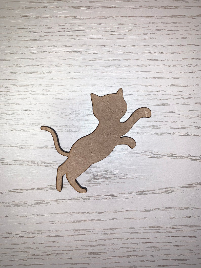 May include: A light brown cat-shaped wooden cutout, mid-jump with one paw extended. The cat has a curved tail and pointed ears. The cutout is set against a light wood-grain background.