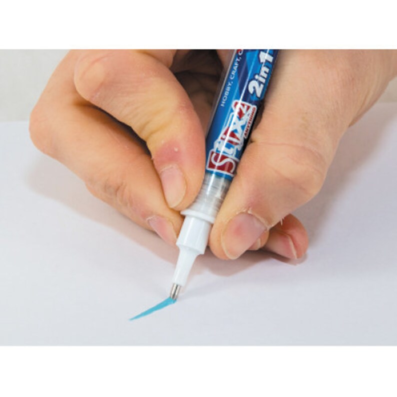 Op de afbeelding: Een blauw-witte Stix 2in1 hobby knutselpen wordt boven een wit oppervlak gehouden, waardoor een dunne, lichtblauwe lijn ontstaat. Op de pen staat de tekst "Stix 2in1" en "Hobby Craft, Anything". De pen heeft een witte punt met een metalen punt.