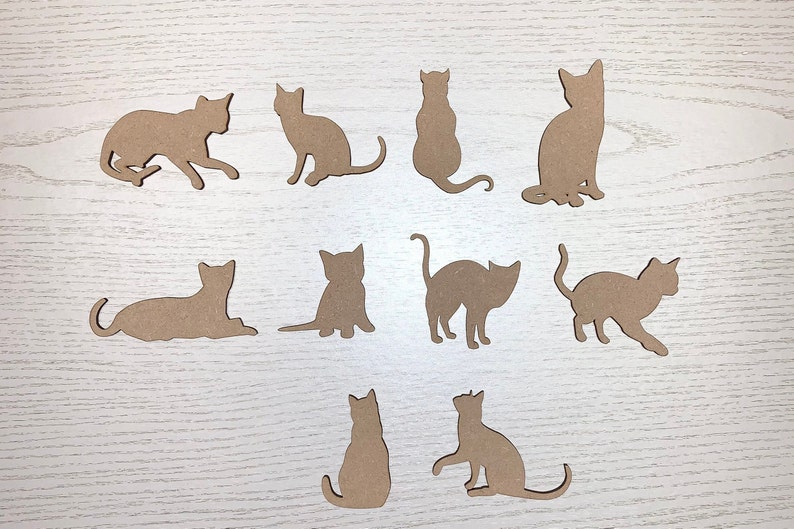 May include: Eleven cat silhouettes in different poses, cut from light brown material. The cats are shown sitting, standing, and walking, with their tails and ears clearly defined. The shapes are arranged on a light wood-grain surface.