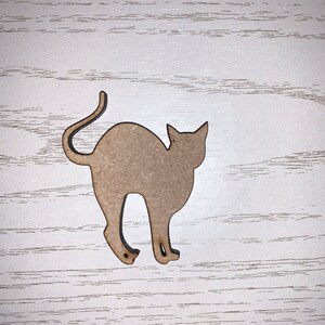 May include: A light brown wooden cat-shaped cutout, featuring a curved tail and pointed ears. The cat is depicted in a walking pose, with its legs slightly bent. The cutout is placed on a white wooden surface.