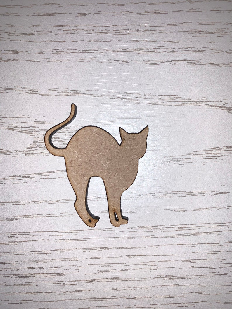 May include: A light brown wooden cat-shaped cutout, featuring a curved tail and pointed ears. The cat is depicted in a walking pose, with its legs slightly bent. The cutout is placed on a white wooden surface.