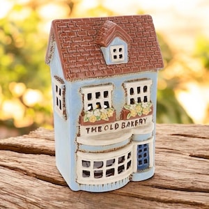 May include: A miniature ceramic house with a blue facade, brown tiled roof, and white window frames. The sign reads "THE OLD BAKERY". The house features a small blue door and is decorated with floral displays.