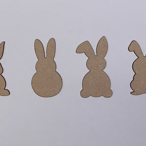 May include: Four wooden bunny shapes, cut out from brown cardboard. The bunnies are all facing forward with their ears up. The bunny on the left has a small flower design on its backside.