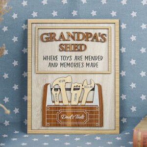Pode incluir: Placa emoldurada de madeira com o texto "GRANDPA'S SHED" e "WHERE TOYS ARE MENDED AND MEMORIES MADE". Abaixo, uma caixa de ferramentas de madeira com ferramentas e o texto "Dad's Tools". Os nomes Peyton, Kevin, Evelyn e Declan também são visíveis.