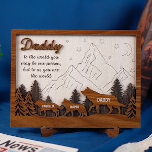 Pode incluir: Um letreiro de madeira com uma cena de montanha e o texto "Daddy to the world you may be one person, but to us you are the world". O letreiro também apresenta três lobos com os nomes "Daddy", "Camellia" e "Jasper" abaixo.