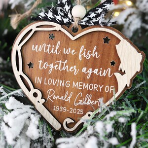 Pode incluir: Enfeite de Natal em forma de coração de madeira com o texto "until we fish together again" e "In loving memory of Ronald Gilbert 1939-2025". O enfeite tem um design de vara de pesca e uma silhueta de uma pessoa pescando. É decorado com um laço preto e branco.
