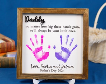 May include: A framed print with a message for a father, "Daddy, no matter how big these hands grow, we'll always be your little ones." The print features two handprints, one pink and one purple, and the text "Love, Devlin and Jessica, Father's Day 2024."