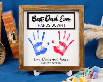 May include: A framed wooden sign with the text "Best Dad Ever Hands Down!" and two handprints in blue and red paint. The sign also includes the text "Love, Devlin and Jessica Father's Day 2024".