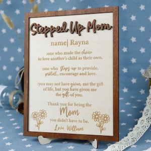 May include: A wooden plaque with a dark brown frame and a light-colored interior. The text "Stepped Up Mom" is at the top, with the name "Rayna" below. The plaque includes a heartfelt message and is supported by a wooden stand.
