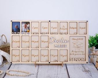May include: A wooden photo frame with 12 slots for pictures, designed to track a child's growth from birth to graduation. The frame is engraved with the child's name, "Devlin Marie", and the graduation year, "Class of 2039". The frame also includes the text "The days are long, but the years are short" and "SENIOR".