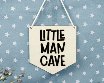 May include: A wooden sign with the words "Little Man Cave" in black lettering. The sign is hanging from a white string against a blue background with white stars.