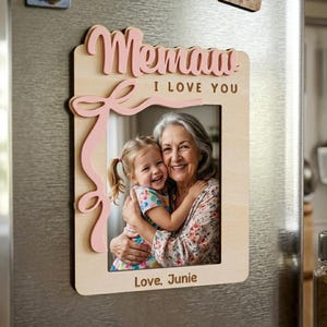 May include: Wooden photo frame with the word "Memaw" in pink script, the phrase "I LOVE YOU", and the words "Love, Junie". The frame holds a photo of a smiling woman and a young girl hugging. The frame is attached to a refrigerator.