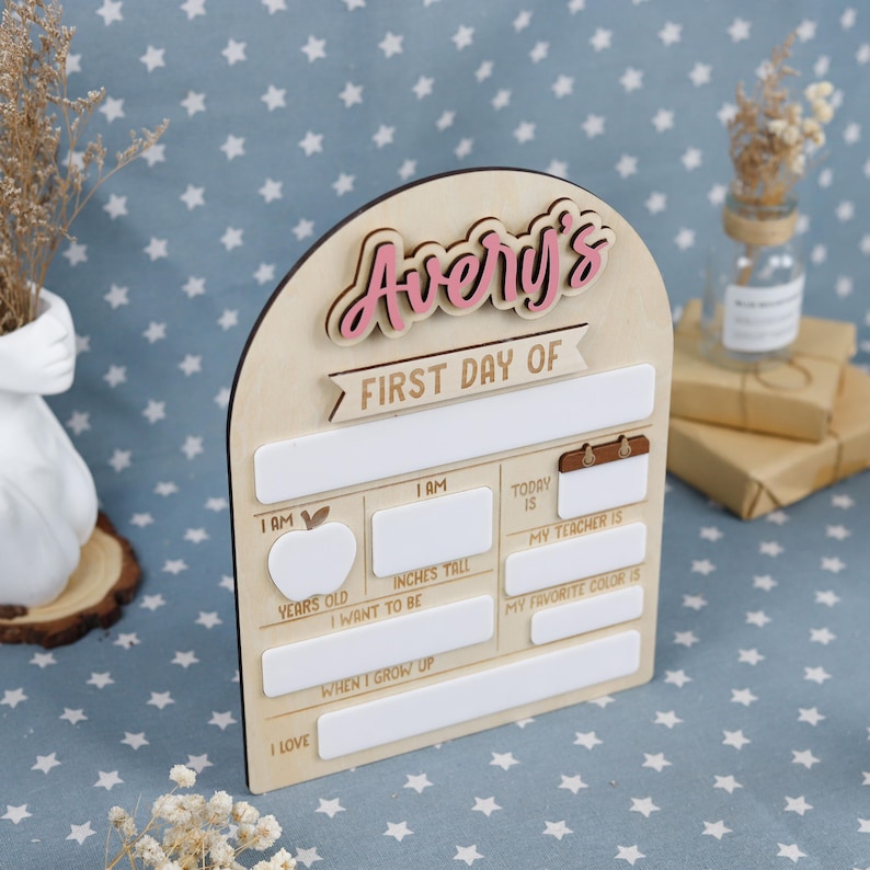 May include: Wooden sign with the text "Avery's First Day Of". The sign has a light wood finish with pink and white accents. It includes spaces to write details about the first day of school, such as age, height, and favorite color.