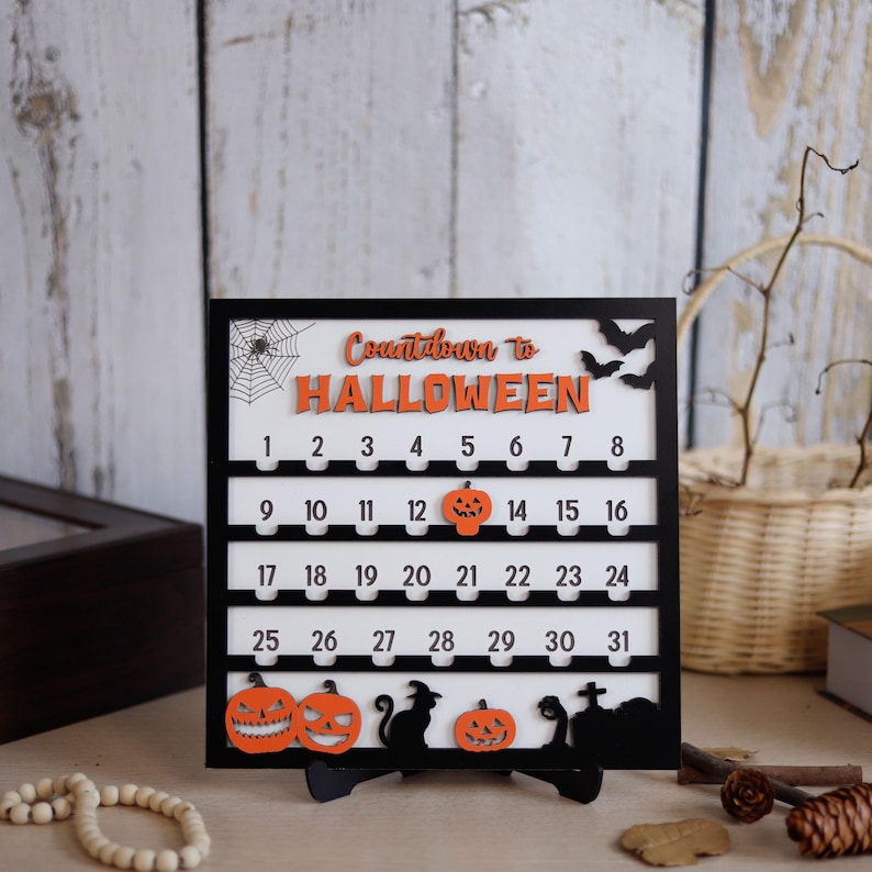 May include: A black framed Halloween countdown calendar with a white background. The calendar features orange text that reads "Countdown to Halloween" and numbers 1-31. Decorative pumpkins, bats, and a spiderweb are also included.