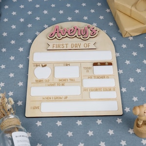 May include: Wooden first day of school sign with the name "Avery's" in pink script. The sign has spaces to fill in information like age, height in inches, teacher's name, and favorite color. The sign is on a blue star-patterned surface.