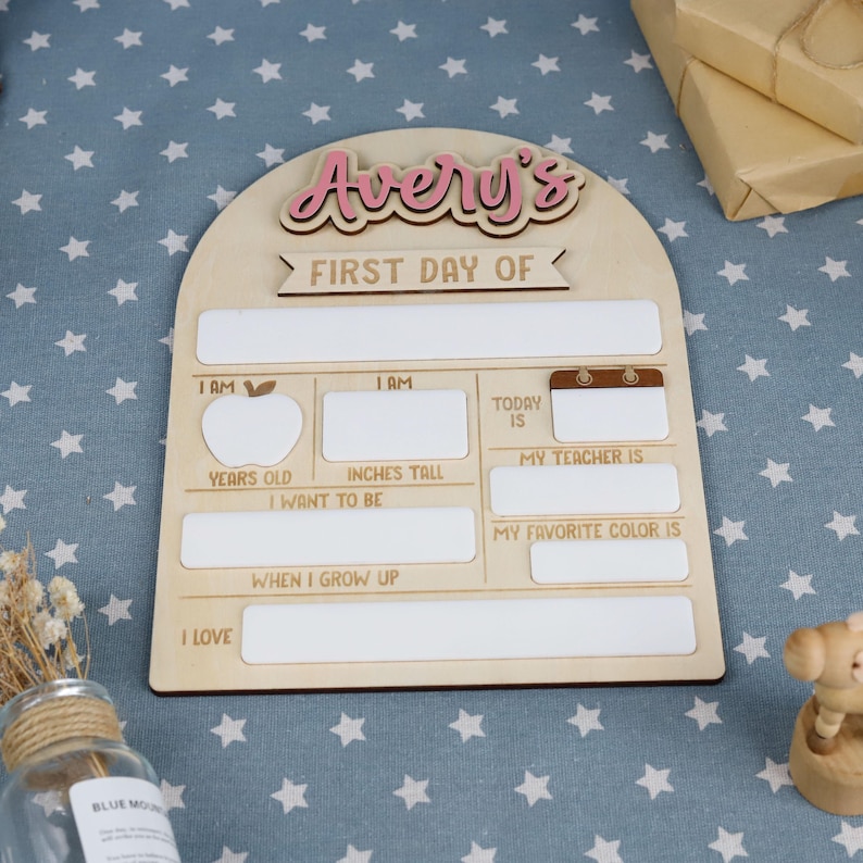 May include: Wooden first day of school sign with the name "Avery's" in pink script. The sign has spaces to fill in information like age, height in inches, teacher's name, and favorite color. The sign is on a blue star-patterned surface.