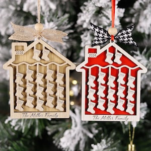 May include: Two wooden house-shaped Christmas ornaments. One is natural wood, the other red. Both have the year 2024, family names, and stockings. The text "The Miller Family" is at the bottom of each ornament.