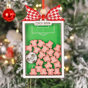 Pode incluir: Enfeite de Natal com tema de futebol. Apresenta um design de campo verde com uma moldura branca, uma bola de futebol e pequenas camisas rosa com n&uacute;meros de jogadores. A parte superior tem um la&ccedil;o vermelho e branco e o texto "COACH BRIAN".