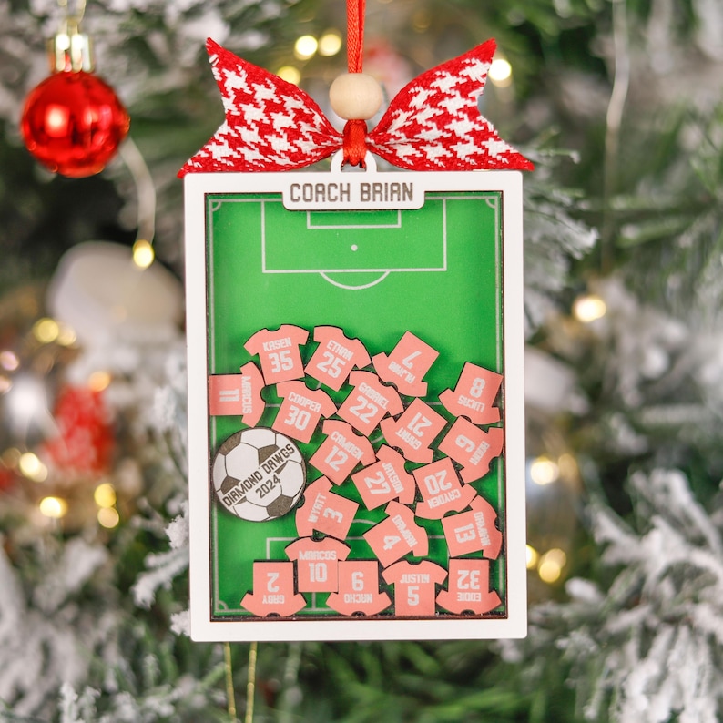 Pode incluir: Enfeite de Natal com tema de futebol. Apresenta um design de campo verde com uma moldura branca, uma bola de futebol e pequenas camisas rosa com n&uacute;meros de jogadores. A parte superior tem um la&ccedil;o vermelho e branco e o texto "COACH BRIAN".