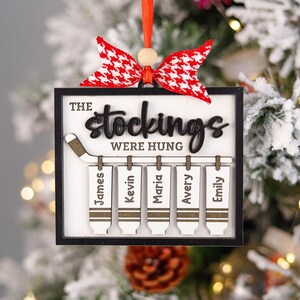 May include: A white wooden Christmas bauble with a black hockey stick and the text "The stockings were hung" with five stockings hanging from the stick, each with a name: James, Kevin, Maria, Avery, and Emily.