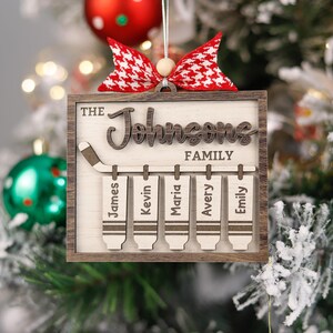 May include: A wooden Christmas bauble featuring a hockey stick and five hanging tags. The bauble is adorned with a red and white checked bow. The text "The Johnsons Family" is inscribed on the bauble. The tags are labelled with the names "James", "Kevin", "Maria", "Avery", and "Emily".