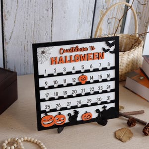 May include: A black and white Halloween countdown calendar with orange accents. The calendar features the text "Countdown to Halloween" and numbers 1-31. Decorative elements include pumpkins, bats, a spiderweb, and tombstones. The calendar is displayed on a wooden surface.