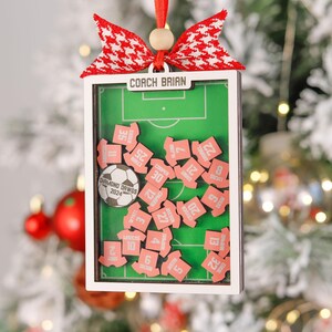 Pode incluir: Enfeite de Natal com tema de futebol. Apresenta um campo de futebol em miniatura com uma bola e camisas de equipe. O texto "COACH BRIAN" &eacute; exibido na parte superior, com o nome da equipe "DIAMOND DAWGS 2024" na bola.