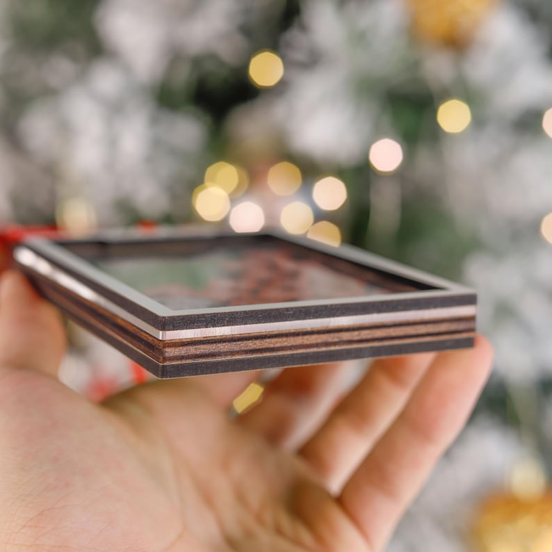 Pode incluir: Um enfeite de Natal quadrado de madeira com um centro transparente. O enfeite tem uma moldura marrom escura com uma borda prateada. As camadas de madeira s&atilde;o vis&iacute;veis na lateral. O fundo est&aacute; desfocado com luzes de Natal.