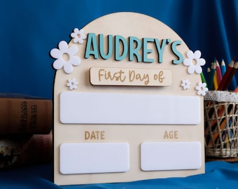 May include: Wooden sign with a light blue arch and the name "Audrey's" in light blue letters. The sign says "First Day of" and has blank spaces for the date and age. The sign is decorated with white flowers.