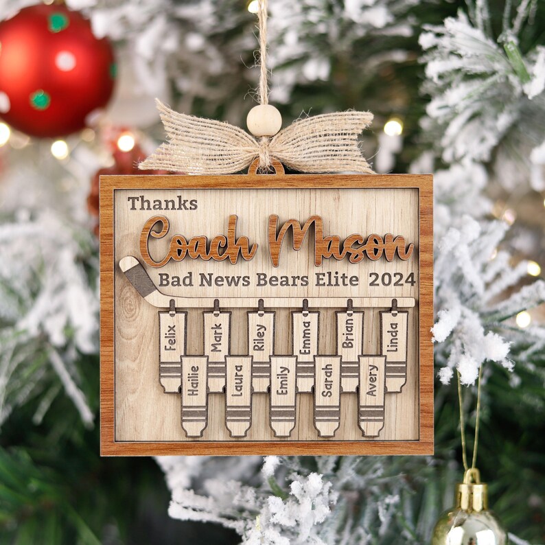 May include: A wooden Christmas bauble featuring a hockey stick and the text "Thanks Coach Mason Bad News Bears Elite 2024". The ornament has a brown background and displays the names of the team members: Felix, Hailie, Mark, Laura, Riley, Emily, Emma, Sarah, Brian, Avery, and Linda.
