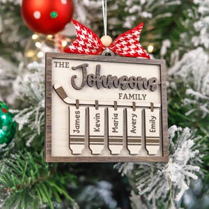 May include: A wooden Christmas bauble featuring a hockey stick and five hockey skate decorations hanging from it. The bauble is personalised with the text "The Johnsons Family" and the names "James, Kevin, Maria, Avery, and Emily".