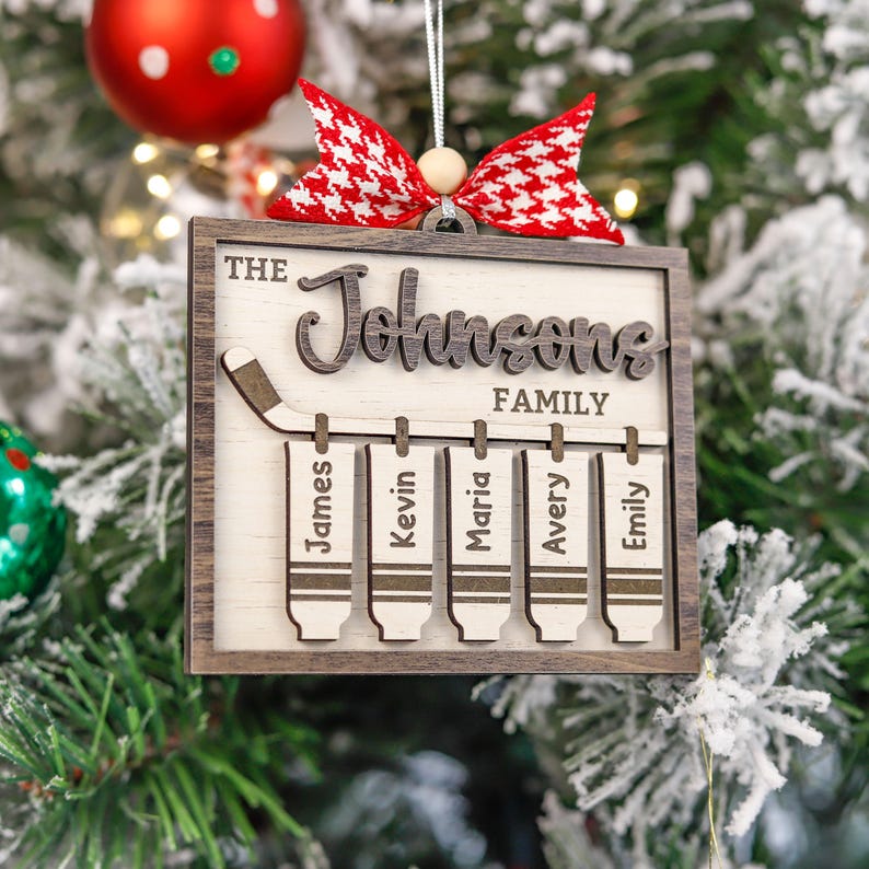 May include: A wooden Christmas bauble featuring a hockey stick and five hockey skate decorations hanging from it. The bauble is personalised with the text "The Johnsons Family" and the names "James, Kevin, Maria, Avery, and Emily".