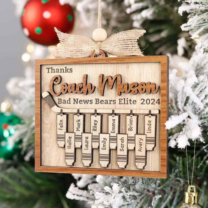 May include: A wooden Christmas bauble with the text "Thanks Coach Mason Bad News Bears Elite 2024". The bauble features a hockey stick with the names of the team members written on individual hockey stick blades.