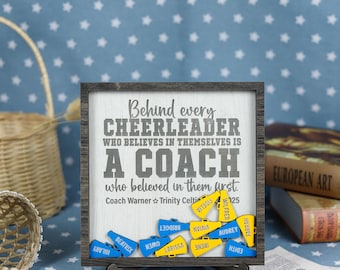 May include: A wooden sign with the text "Behind every cheerleader who believes in themselves is a coach who believed in them first."  The sign has a gray background and white text.  There are small wooden cheerleading megaphones with names written on them in front of the sign.