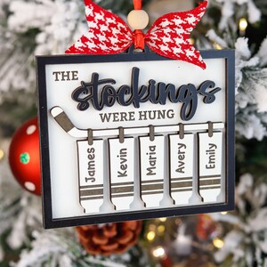 May include: A white wooden Christmas bauble with a black hockey stick and five stockings hanging from it. The text "The Stockings Were Hung" is written on the bauble. The stockings are labelled with the names "James", "Kevin", "Maria", "Avery", and "Emily".