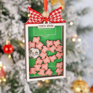 Pode incluir: Enfeite de Natal retangular com um design de campo de futebol. O enfeite apresenta um campo verde, pe&ccedil;as rosa em forma de camisa com n&uacute;meros e uma bola de futebol. O texto "COACH BRIAN" est&aacute; no topo. Uma fita vermelha e branca e uma fita vermelha est&atilde;o presas.