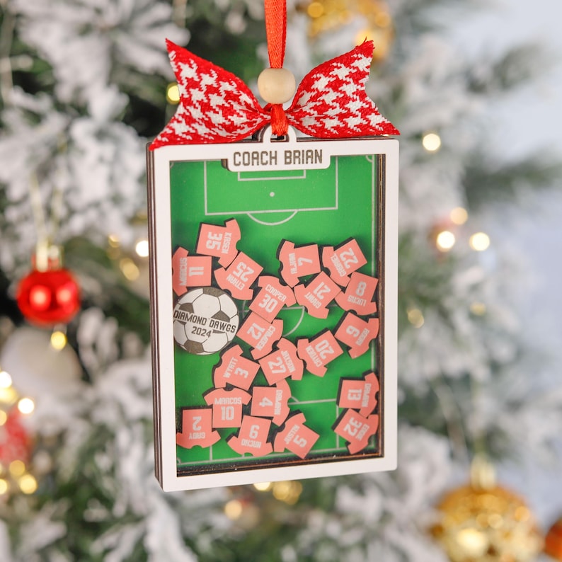 Pode incluir: Enfeite de Natal retangular com um design de campo de futebol. O enfeite apresenta um campo verde, pe&ccedil;as rosa em forma de camisa com n&uacute;meros e uma bola de futebol. O texto "COACH BRIAN" est&aacute; no topo. Uma fita vermelha e branca e uma fita vermelha est&atilde;o presas.
