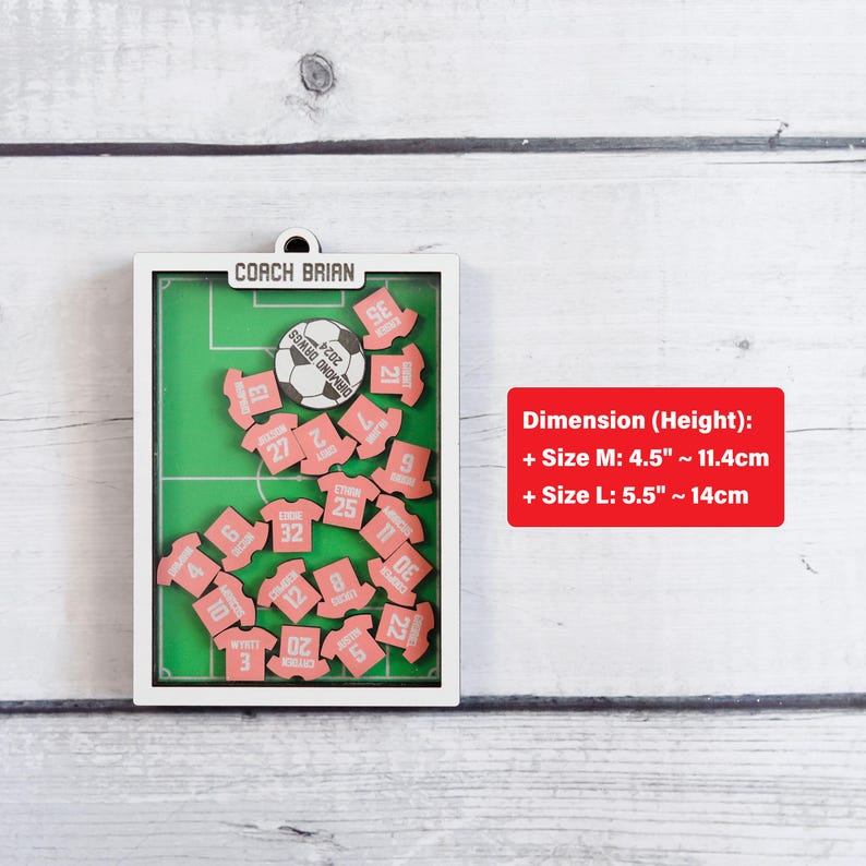 Pode incluir: Um quadro de exibi&ccedil;&atilde;o de madeira com tema de futebol com um design de campo verde. Apresenta uma bola de futebol e pequenas pe&ccedil;as rosa em forma de camisa com n&uacute;meros. O texto "COACH BRIAN" est&aacute; no topo. O tamanho M tem 11,4 cm de altura e o tamanho L tem 14 cm de altura.