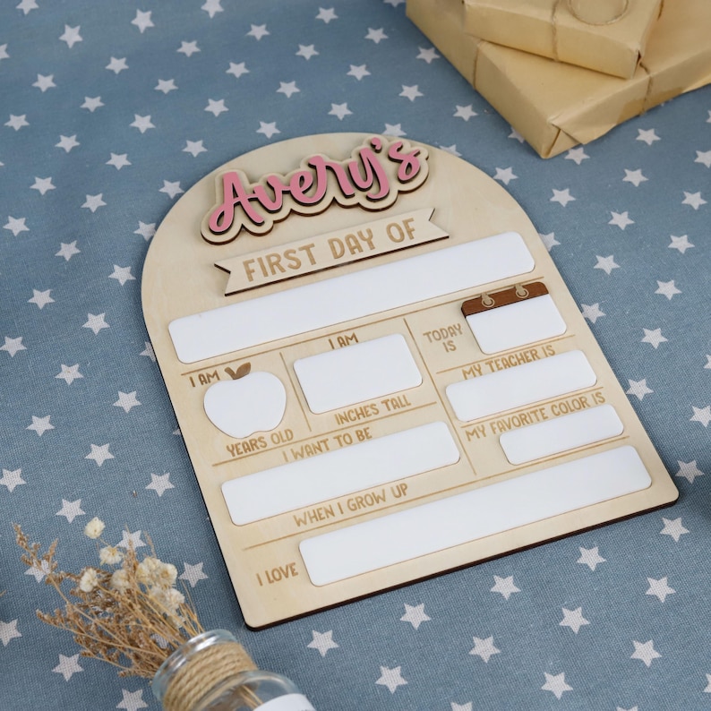 May include: A wooden sign with the text "Avery's FIRST DAY OF" in pink and brown. The sign has spaces to fill in information like age, height, teacher, and favorite color. The sign is on a blue surface with white stars.