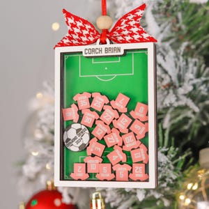 Pode incluir: Enfeite de Natal personalizado com tema de futebol. O enfeite apresenta um campo de futebol em miniatura com etiquetas de nome em forma de camisa rosa e uma bola de futebol. A parte superior do enfeite diz "COACH BRIAN" e &eacute; adornada com um la&ccedil;o vermelho e branco.