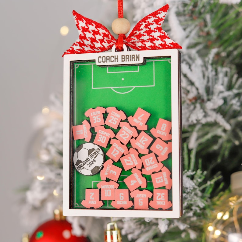 Pode incluir: Enfeite de Natal personalizado com tema de futebol. O enfeite apresenta um campo de futebol em miniatura com etiquetas de nome em forma de camisa rosa e uma bola de futebol. A parte superior do enfeite diz "COACH BRIAN" e &eacute; adornada com um la&ccedil;o vermelho e branco.