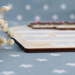 May include: Wooden memory board with a dark brown edge, featuring white rectangular and oval-shaped spaces. The words "I LOVE" are engraved on the board. A pink and brown wooden piece sits on top. Dried white flowers are in the foreground.