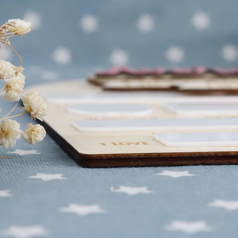 May include: Wooden memory board with a dark brown edge, featuring white rectangular and oval-shaped spaces. The words "I LOVE" are engraved on the board. A pink and brown wooden piece sits on top. Dried white flowers are in the foreground.