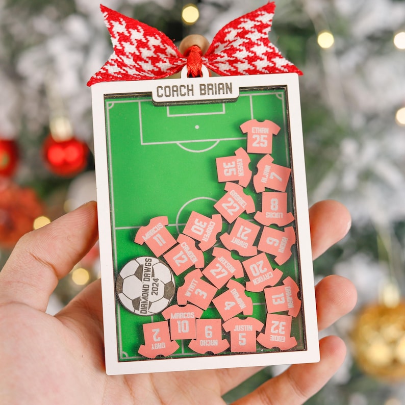 Pode incluir: Um enfeite de futebol com moldura branca com um design de campo verde, com o texto "COACH BRIAN". O enfeite inclui pe&ccedil;as em forma de camisa rosa com nomes e n&uacute;meros de jogadores, uma bola de futebol e um la&ccedil;o vermelho e branco.