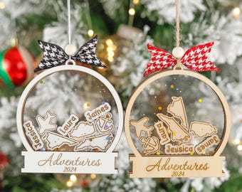 May include: Two personalized Christmas ornaments with clear globes, wooden frames, and decorative bows. Each ornament features names, map shapes, and the word "Adventures" with the year 2024. One bow is black and white, the other is red and white.