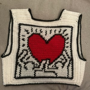 May include: White crocheted sleeveless vest with a black border and a red heart design in the center.