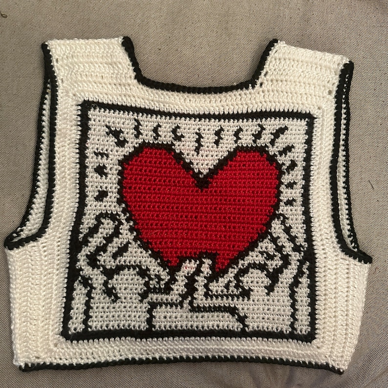 May include: White crocheted sleeveless vest with a black border and a red heart design in the center.