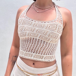 Könnte beinhalten: Beige-weißes Häkeltop mit geometrischem Muster. Das Top hat einen Neckholder-Ausschnitt und eine verkürzte Länge. Es wird mit einer goldfarbenen, perlenbesetzten Taillenkette und hellen Hosen kombiniert. Der Hintergrund ist eine schlichte, weiße Wand.